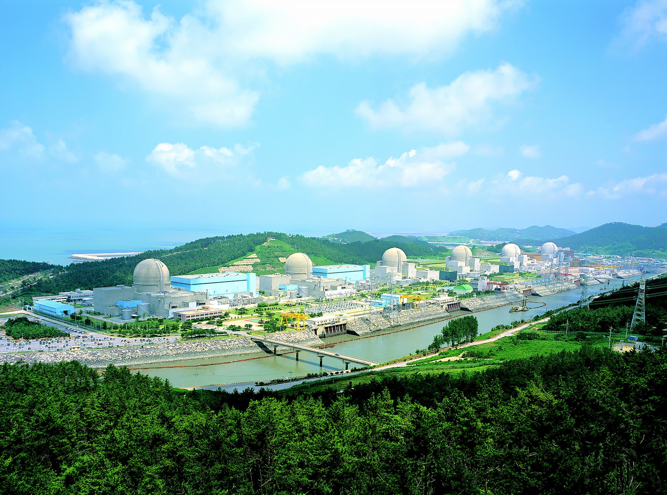 Coastal nuclear power plant with several dome reactors along a river and green hills.